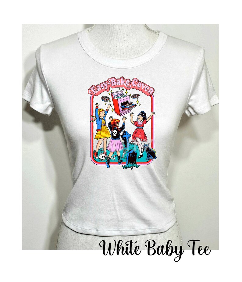 May include: White baby tee shirt with a graphic print of three girls in front of a pink Easy-Bake Oven. The girls are wearing colorful clothing and are holding up the oven. The graphic is surrounded by a red border and the text "Easy-Bake Coven" is written in pink letters.