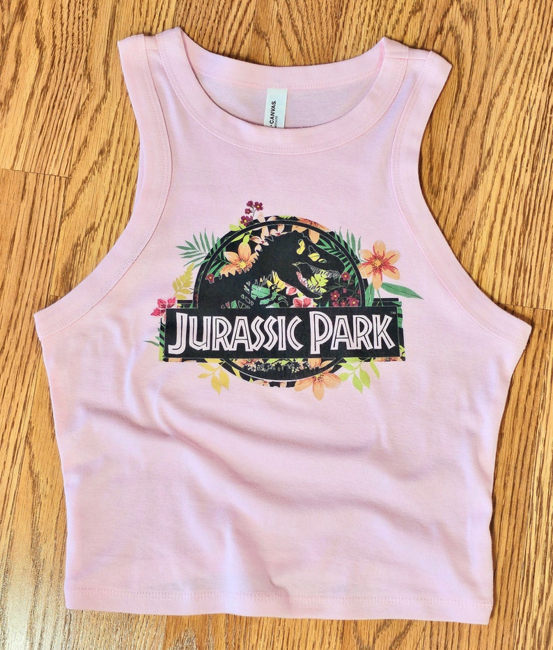 May include: A light pink sleeveless crop top featuring a graphic print of a black dinosaur silhouette inside a circular frame surrounded by colourful tropical flowers and leaves, with the text JURASSIC PARK printed in white on a black rectangular bar.