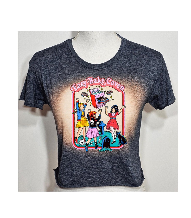 May include: A dark gray cropped t-shirt with a bleached effect. The shirt features a graphic of three girls in front of an Easy-Bake Oven with the text "Easy-Bake Coven".