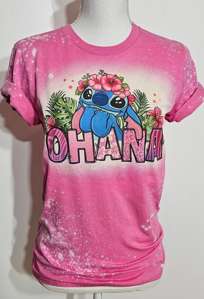 May include: Pink t-shirt with a bleach-splattered design featuring Stitch from Lilo & Stitch wearing a flower crown and the word "Ohana" in colorful letters.