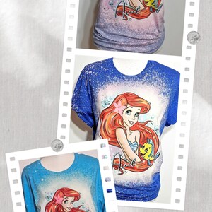 Puede incluir: Una camiseta tie-dye azul, blanca y rosa con un dise&ntilde;o salpicado de lej&iacute;a que presenta a Ariel de La Sirenita sosteniendo a Flounder. La camiseta tiene el texto "Someday I'll be part of your world" y "Ariel" escrito en ella.