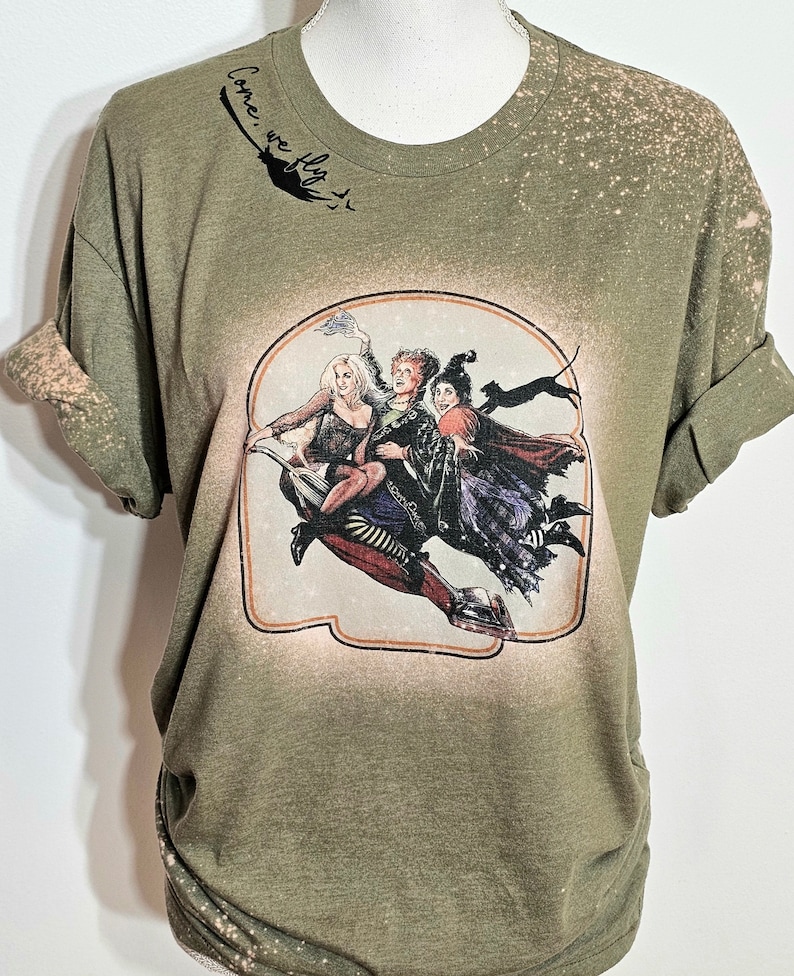 Puede incluir: Camiseta verde con efecto blanqueado y un estampado gr&aacute;fico de tres brujas volando en una escoba. El texto "Come, we fly" est&aacute; escrito sobre el gr&aacute;fico.