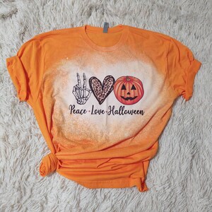 Puede incluir: Camiseta naranja con un dise&ntilde;o blanqueado que presenta un signo de paz, un coraz&oacute;n y una calabaza con el texto "Peace Love Halloween".