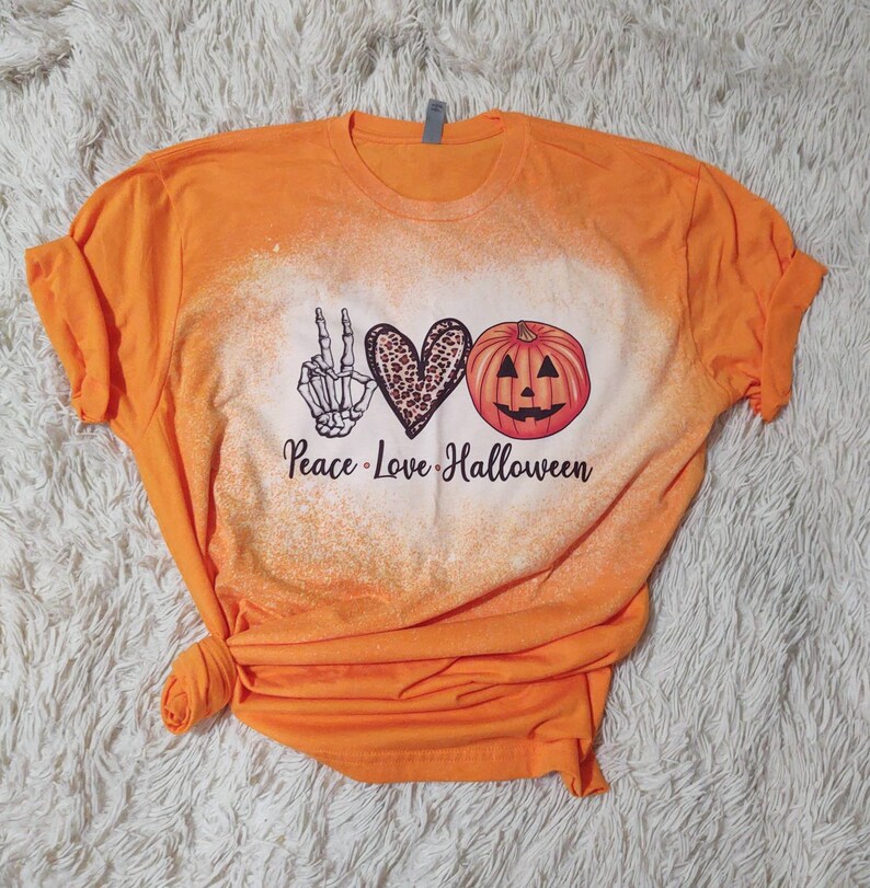 Puede incluir: Camiseta naranja con un dise&ntilde;o blanqueado que presenta un signo de paz, un coraz&oacute;n y una calabaza con el texto "Peace Love Halloween".