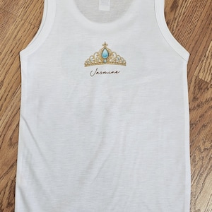 Peut inclure: D&eacute;bardeur blanc avec un motif de couronne dor&eacute;e et turquoise et le nom "Jasmine" imprim&eacute; en dessous. La couronne pr&eacute;sente une grosse gemme turquoise au centre et de plus petites gemmes sur les bords. Le d&eacute;bardeur est en mati&egrave;re l&eacute;g&egrave;re.