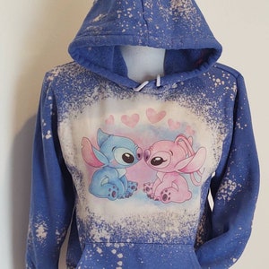 May include: Blue and pink bleached hoodie with a graphic of Stitch and Angel from Lilo & Stitch kissing. The hoodie has a distressed look with white bleach splatters.