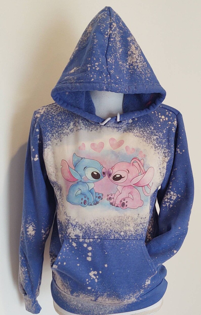 May include: Blue and pink bleached hoodie with a graphic of Stitch and Angel from Lilo & Stitch kissing. The hoodie has a distressed look with white bleach splatters.