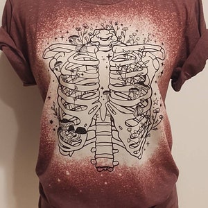 以下が含まれることがあります： マロン色のTシャツに、漂白されたデザインが施されています。肋骨の中に花とキノコが詰まっています。デザインは白黒です。