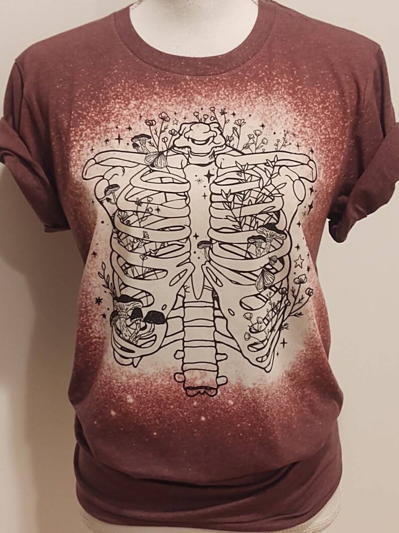 以下が含まれることがあります： マロン色のTシャツに、漂白されたデザインが施されています。肋骨の中に花とキノコが詰まっています。デザインは白黒です。