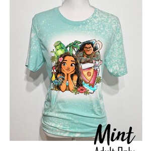 May include: A mint green t-shirt with a bleach-splattered design featuring a cartoon image of Moana, Maui, Pua, and Hei Hei from the Disney movie Moana. The text "Mint Adult Only" is printed below the image.