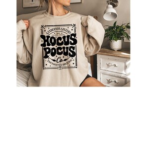May include: A beige sweatshirt with a black graphic print that reads "Sanderson Sisters 1693 Salem, Mass Hocus Pocus Co. Enchanted Brooms".