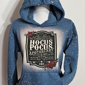 Puede incluir: Una sudadera con capucha azul con un dise&ntilde;o salpicado de lej&iacute;a blanca. La parte delantera de la sudadera presenta un gr&aacute;fico en blanco y negro de una etiqueta que dice "The Sanderson Sisters Hocus Pocus Apothecary" con el texto "Fine Potions, Magic Elixirs, Mystical Brews, Tonics & Tinctures" debajo. La etiqueta est&aacute; rodeada de rosas rojas.