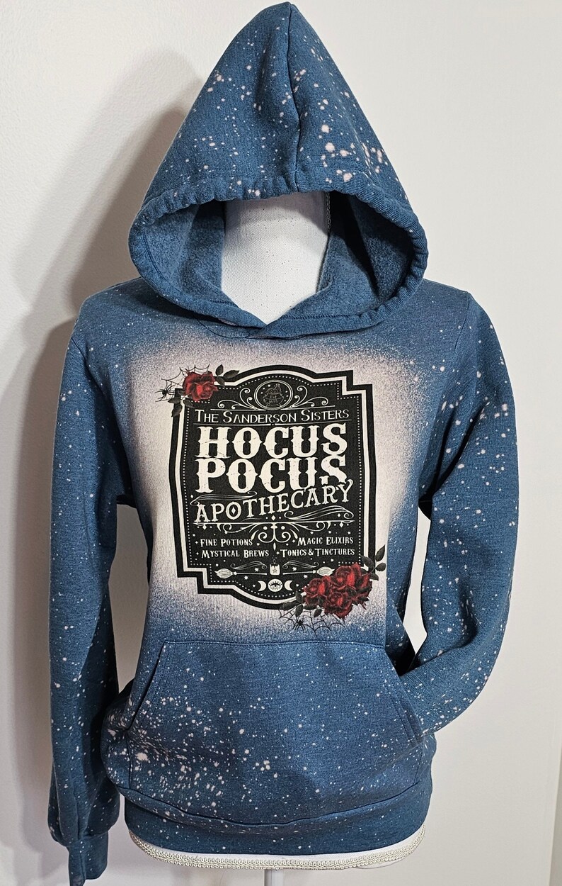 Puede incluir: Una sudadera con capucha azul con un dise&ntilde;o salpicado de lej&iacute;a blanca. La parte delantera de la sudadera presenta un gr&aacute;fico en blanco y negro de una etiqueta que dice "The Sanderson Sisters Hocus Pocus Apothecary" con el texto "Fine Potions, Magic Elixirs, Mystical Brews, Tonics & Tinctures" debajo. La etiqueta est&aacute; rodeada de rosas rojas.