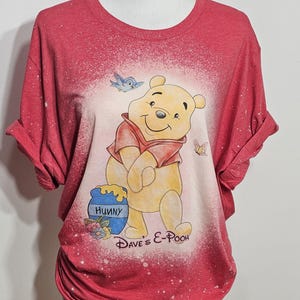 May include: A red short-sleeve t-shirt with a bleached effect featuring a graphic of Winnie the Pooh, a blue bird, a butterfly, and a honey pot labeled "Hunny". The text "Dave's E-Pooh" is printed in black script below the character illustration.