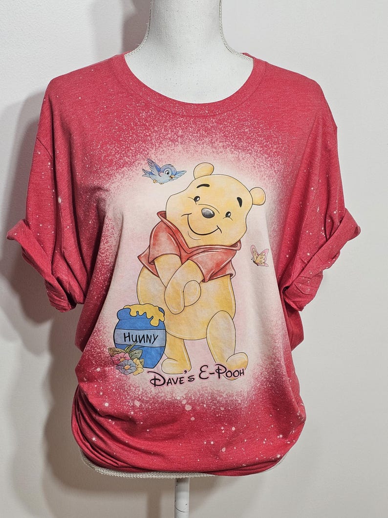 May include: A red short-sleeve t-shirt with a bleached effect featuring a graphic of Winnie the Pooh, a blue bird, a butterfly, and a honey pot labeled "Hunny". The text "Dave's E-Pooh" is printed in black script below the character illustration.