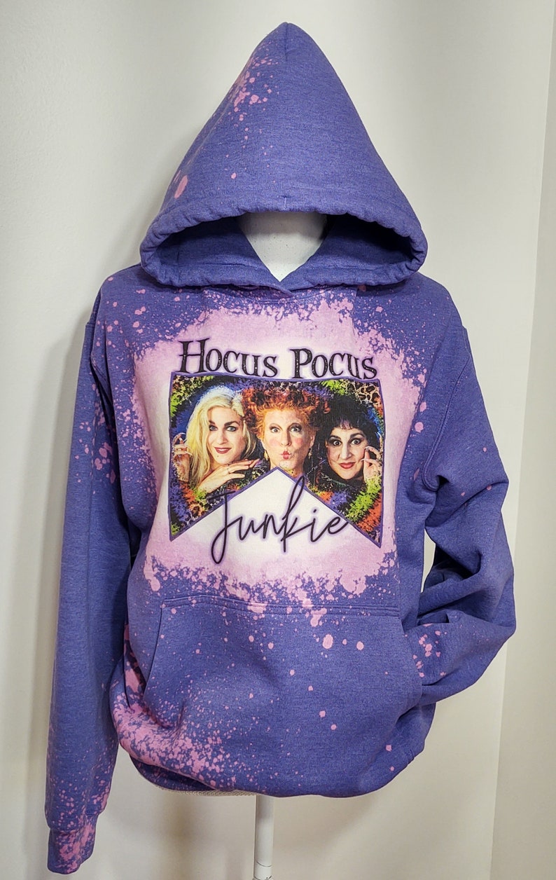 May include: Purple hooded sweatshirt with a bleach-splattered design featuring a picture of the Sanderson sisters from the movie Hocus Pocus and the text "Hocus Pocus Junkie".