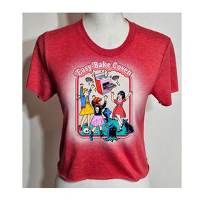 May include: Red cropped t-shirt with a graphic print of three girls in front of a tombstone and an Easy-Bake Oven. The text "Easy-Bake Coven" is printed above the graphic.