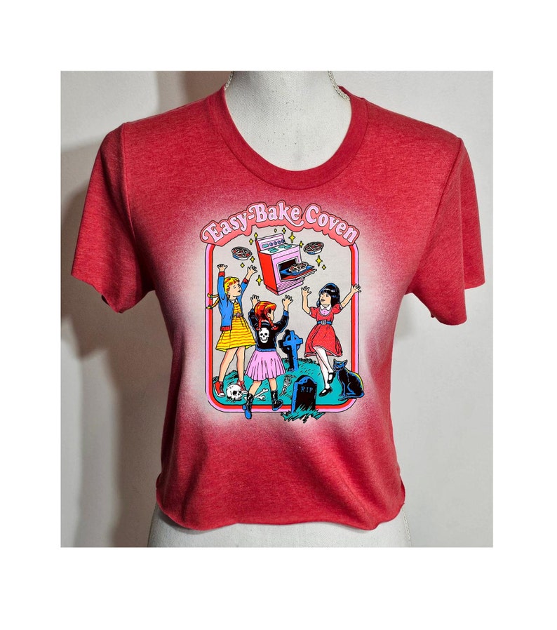 May include: Red cropped t-shirt with a graphic print of three girls in front of a tombstone and an Easy-Bake Oven. The text "Easy-Bake Coven" is printed above the graphic.