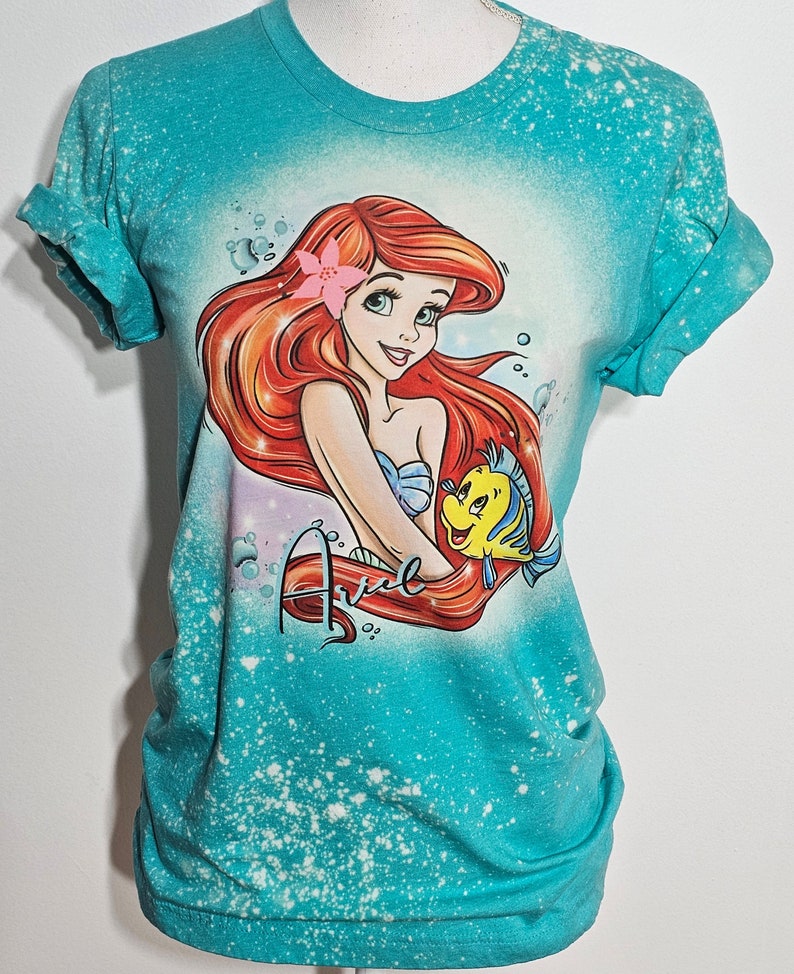 Puede incluir: Una camiseta azul turquesa con un dise&ntilde;o salpicado de lej&iacute;a que presenta a Ariel, la Sirenita, con su cabello rojo y una flor rosa en el cabello. Ella est&aacute; sosteniendo un pez amarillo con una aleta caudal azul. La palabra "Ariel" est&aacute; escrita en cursiva roja debajo de ella.