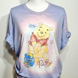 May include: A light blue bleached t-shirt featuring a graphic of Winnie the Pooh in a red shirt, a blue honey pot labeled Hunny, a blue bird, and a butterfly. The text Pooh Bear is printed in a cursive script below the character illustration.