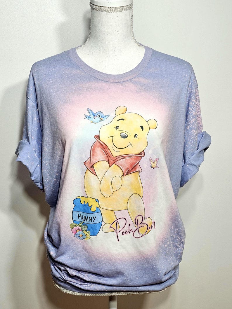May include: A light blue bleached t-shirt featuring a graphic of Winnie the Pooh in a red shirt, a blue honey pot labeled Hunny, a blue bird, and a butterfly. The text Pooh Bear is printed in a cursive script below the character illustration.