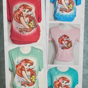 Puede incluir: Cinco camisetas de diferentes colores con un dise&ntilde;o salpicado de lej&iacute;a con una imagen de dibujos animados de Ariel de La Sirenita sosteniendo un pez amarillo. Las camisetas son de color rojo, azul, rosa, verde y turquesa.