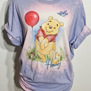 May include: A light purple bleached t-shirt featuring a graphic of Winnie the Pooh holding a red balloon with a blue bird flying above. The design includes green grass, a small mushroom, and the word Honey written in a blue cursive script font.