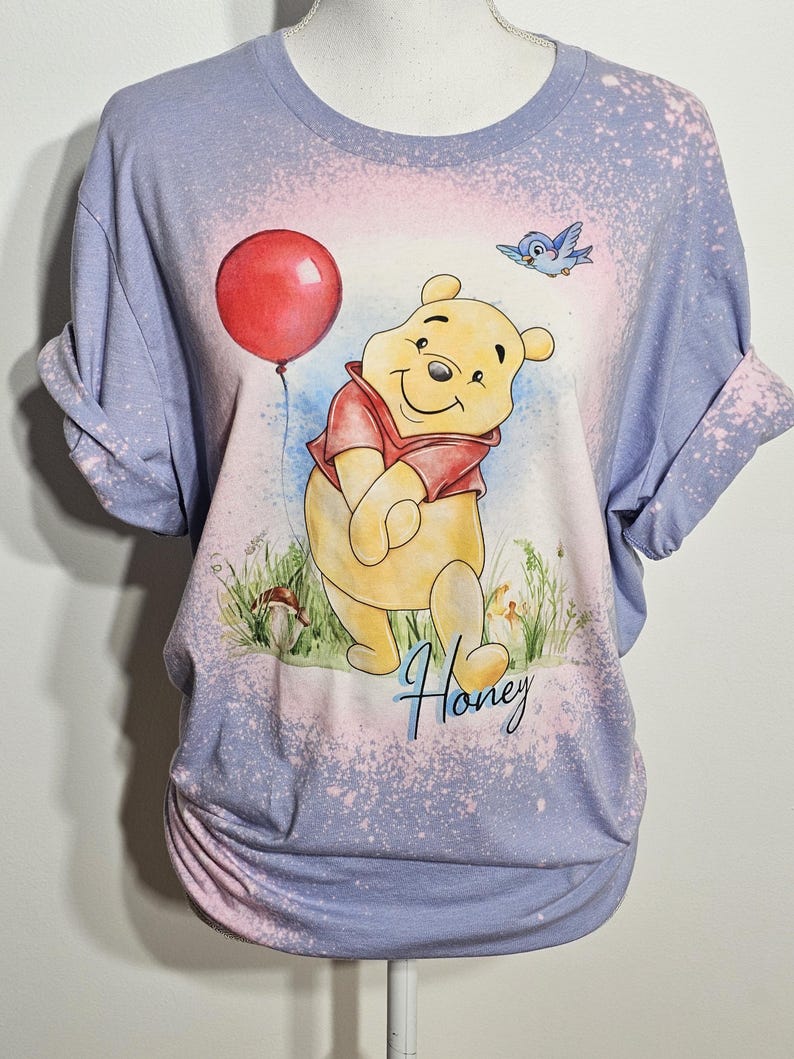 May include: A light purple bleached t-shirt featuring a graphic of Winnie the Pooh holding a red balloon with a blue bird flying above. The design includes green grass, a small mushroom, and the word Honey written in a blue cursive script font.