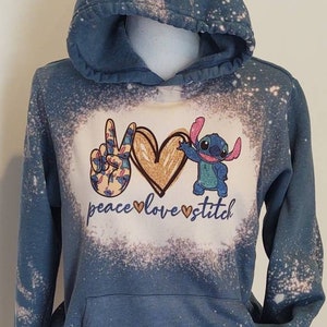 May include: A blue hooded sweatshirt with a bleach-splattered design. The front of the sweatshirt features a peace sign, a heart, and a cartoon Stitch character. The text "peace love stitch" is printed below the design.