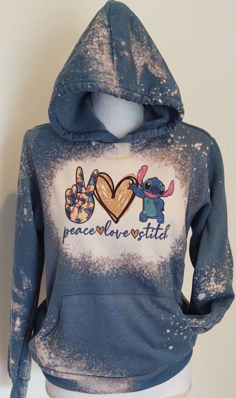 May include: A blue hooded sweatshirt with a bleach-splattered design. The front of the sweatshirt features a peace sign, a heart, and a cartoon Stitch character. The text "peace love stitch" is printed below the design.