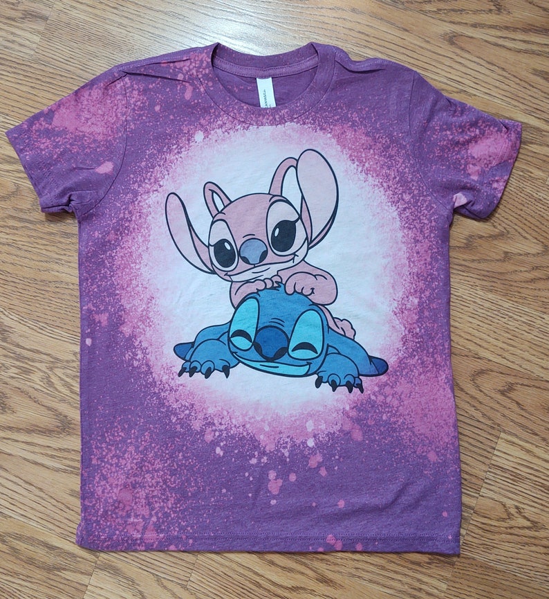 May include: Purple tie-dye t-shirt with a white graphic of Stitch and Angel from the Disney movie Lilo & Stitch.