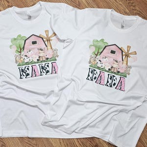 May include: Two white t-shirts with a farm-themed graphic. The design features a pink barn, windmill, and various farm animals. The word "MAMA" is printed in a cow-print and pink plaid font.
