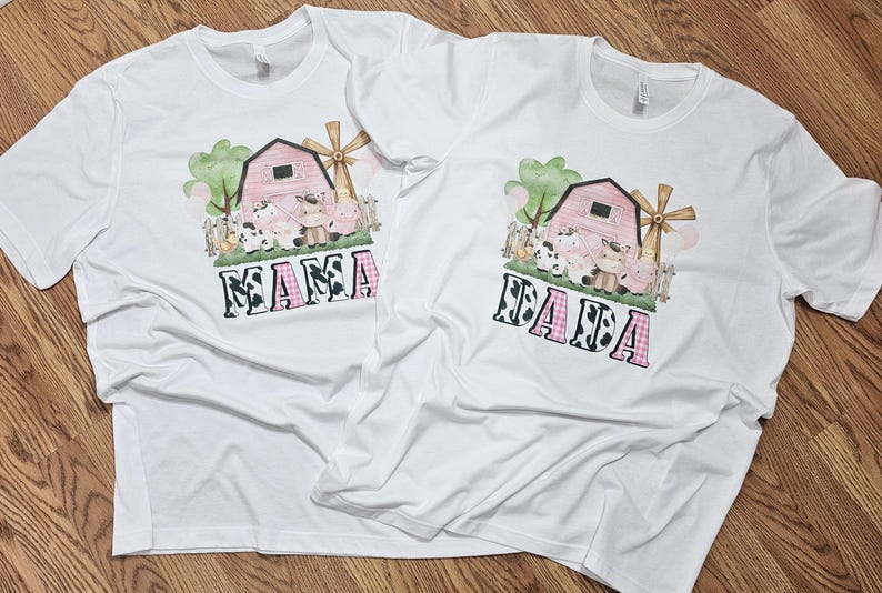 May include: Two white t-shirts with a farm-themed graphic. The design features a pink barn, windmill, and various farm animals. The word "MAMA" is printed in a cow-print and pink plaid font.