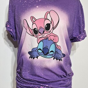 May include: Purple t-shirt with a bleach-splattered design featuring Stitch and Angel from Lilo & Stitch. The characters are pink and blue and are smiling.