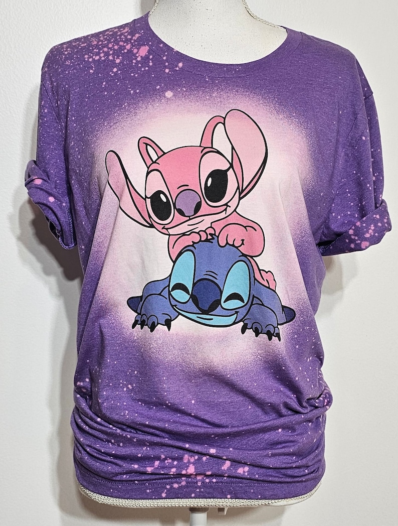 May include: Purple t-shirt with a bleach-splattered design featuring Stitch and Angel from Lilo & Stitch. The characters are pink and blue and are smiling.