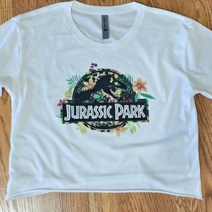 May include: A white short-sleeved crop top featuring a graphic print of the Jurassic Park logo in black, surrounded by colourful tropical flowers and green leaves. The text Jurassic Park is printed in white letters on a black rectangular background.