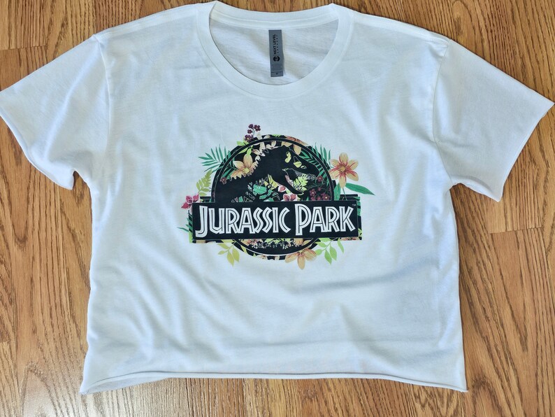 May include: A white short-sleeved crop top featuring a graphic print of the Jurassic Park logo in black, surrounded by colourful tropical flowers and green leaves. The text Jurassic Park is printed in white letters on a black rectangular background.