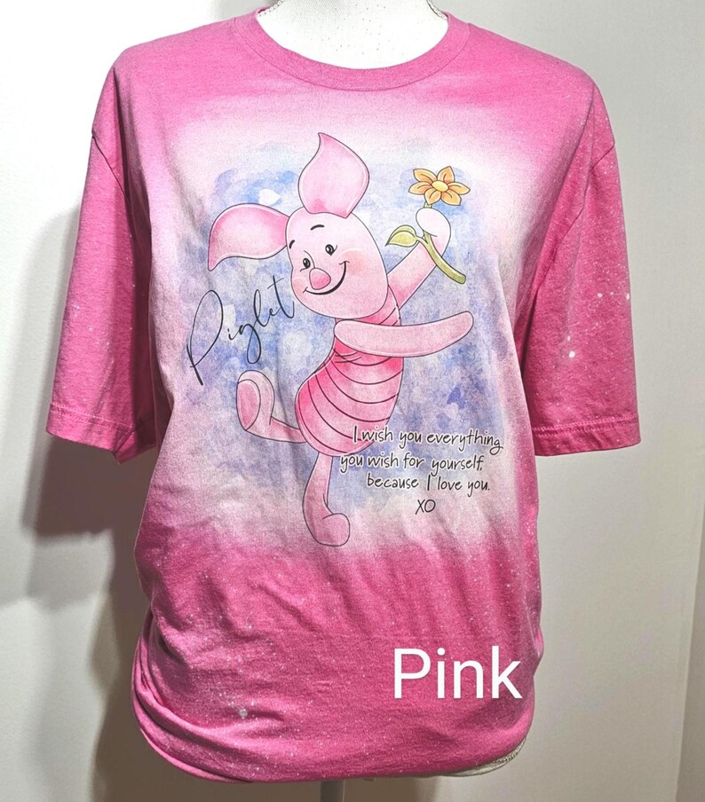 May include: A pink bleached t-shirt featuring a graphic of Piglet holding a yellow flower. The text reads Piglet and I wish you everything you wish for yourself because I love you. XO. The shirt has a faded pink color with white bleached spots.