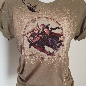 May include: A green t-shirt with a bleach-splattered design featuring a vintage-style image of three witches flying on a broomstick. The text "Come, we fly" is written in cursive on the top of the shirt.