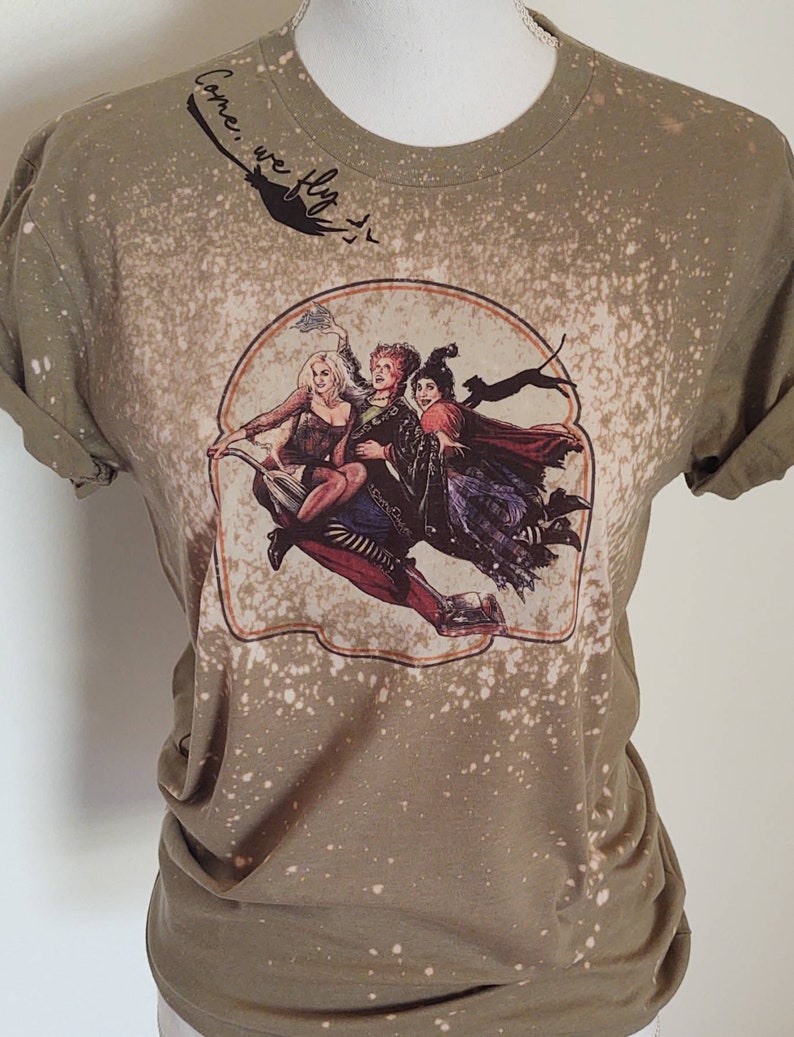 May include: A green t-shirt with a bleach-splattered design featuring a vintage-style image of three witches flying on a broomstick. The text "Come, we fly" is written in cursive on the top of the shirt.