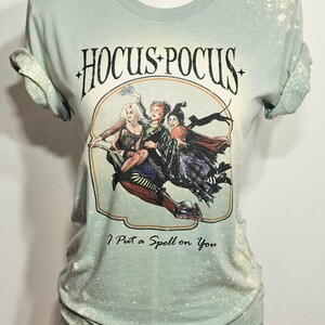 Puede incluir: Una camiseta verde con efecto blanqueado que presenta un gr&aacute;fico de las hermanas Sanderson de la pel&iacute;cula "Hocus Pocus" volando en una escoba. El texto "Hocus Pocus" y "I Put a Spell on You" est&aacute; impreso en la camiseta.