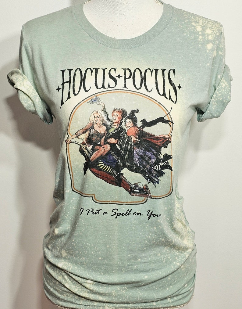 Puede incluir: Una camiseta verde con efecto blanqueado que presenta un gr&aacute;fico de las hermanas Sanderson de la pel&iacute;cula "Hocus Pocus" volando en una escoba. El texto "Hocus Pocus" y "I Put a Spell on You" est&aacute; impreso en la camiseta.