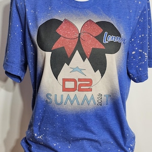 May include: Blue t-shirt with a graphic featuring Mickey Mouse ears, a red bow, and the name "Lennox." Below the ears is the text "D2 SUMMIT 2026" in white. The shirt has a speckled design.