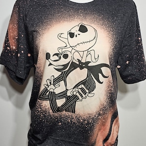 以下が含まれることがあります： ナイトメア・ビフォア・クリスマスのジャック・スケリントンとゼロの白いシルエットが描かれた、漂白剤で飛び散ったデザインの黒いTシャツです。