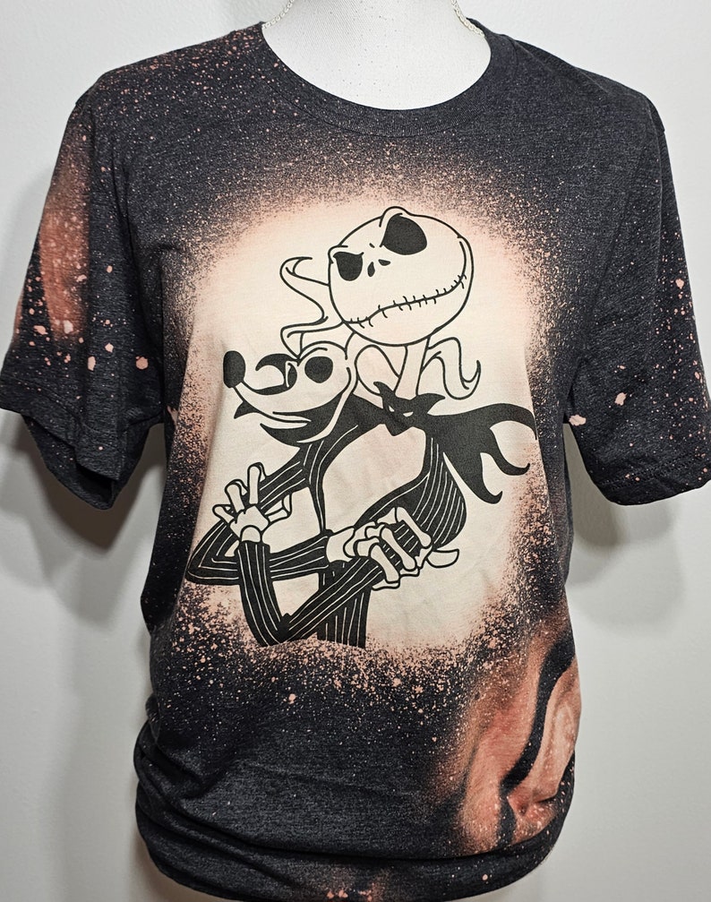 以下が含まれることがあります： ナイトメア・ビフォア・クリスマスのジャック・スケリントンとゼロの白いシルエットが描かれた、漂白剤で飛び散ったデザインの黒いTシャツです。