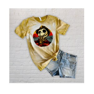 以下が含まれることがあります： 帽子をかぶってナイフを持ったスケルトンの円形のグラフィックがプリントされた、黄色の漂白されたTシャツ。グラフィックは、木のある夕焼けの背景に設定されています。