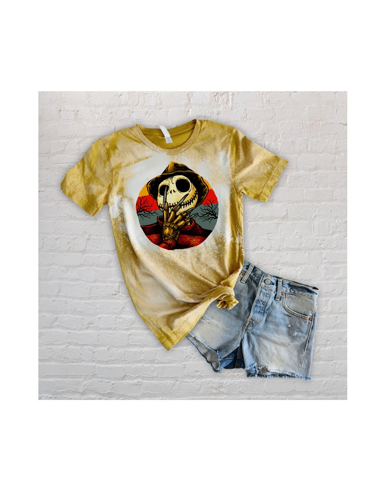 以下が含まれることがあります： 帽子をかぶってナイフを持ったスケルトンの円形のグラフィックがプリントされた、黄色の漂白されたTシャツ。グラフィックは、木のある夕焼けの背景に設定されています。