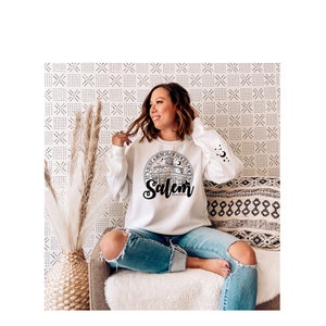 May include: A white sweatshirt with a black graphic print of a building with the text "It's just a bunch of hocus pocus Salem, Massachusetts" on a white background. The model is wearing blue jeans and sitting on a couch.