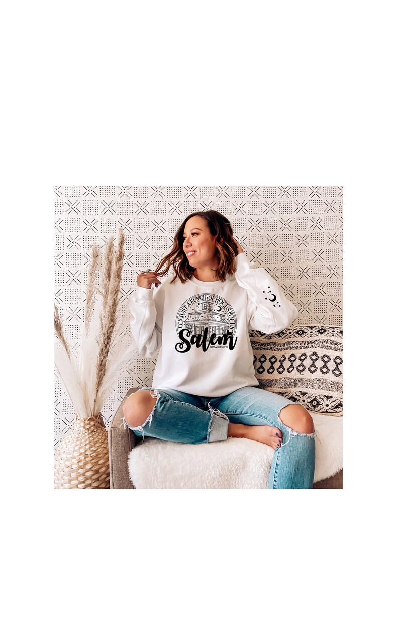 May include: A white sweatshirt with a black graphic print of a building with the text "It's just a bunch of hocus pocus Salem, Massachusetts" on a white background. The model is wearing blue jeans and sitting on a couch.