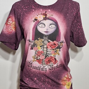以下が含まれることがあります： 白とピンクの漂白剤でデザインされた、マロン色のTシャツ。骸骨に花が飾られ、「Beautifully Broken」という文字が書かれている。